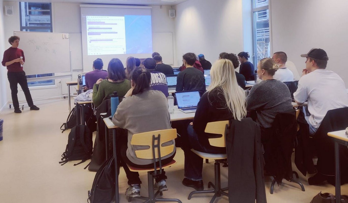 Students sitting in the classroom and facing the front of the room where a teacher is standing next to a whiteboard with some information written on it. On a screen projected image of a website decoloniale. 